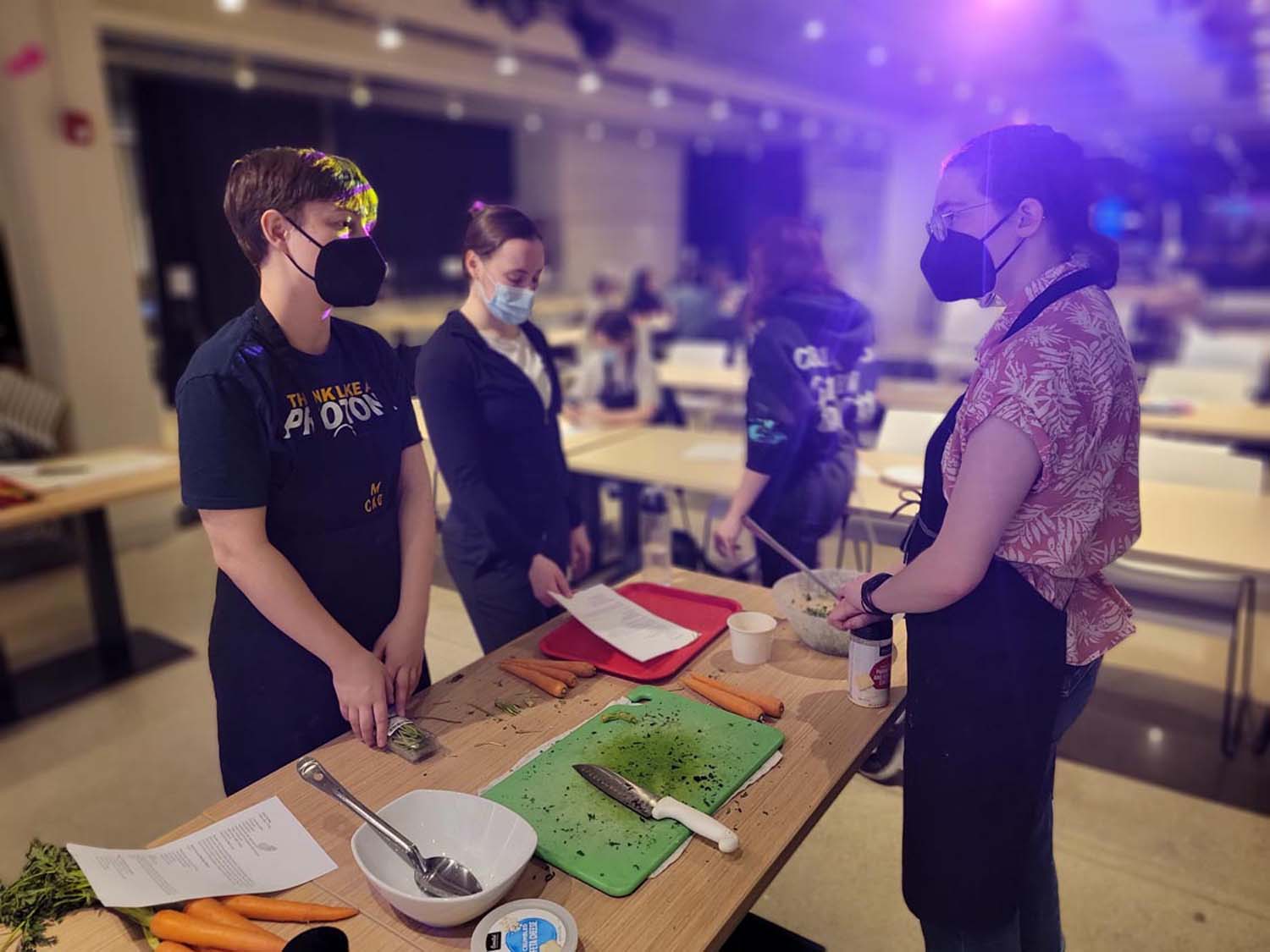 Three people stand around a table with cooking tools and ingredients on it 