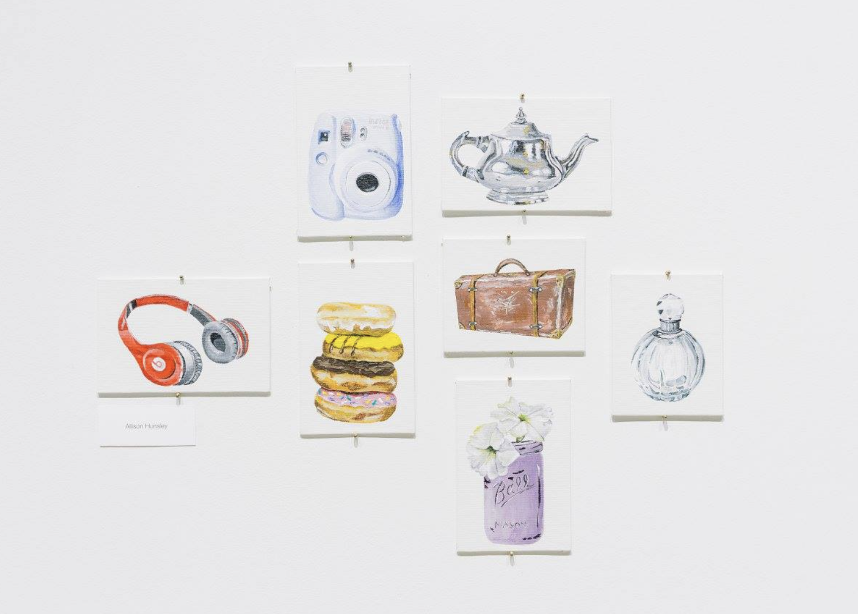 Painting student work featuring seven small paintings on a white wall. Objects in paintings include headphones, a stack of doughnuts, a vintage suitcase, a perfume bottle, a teapot, a jar of flowers, and a poloroid camera