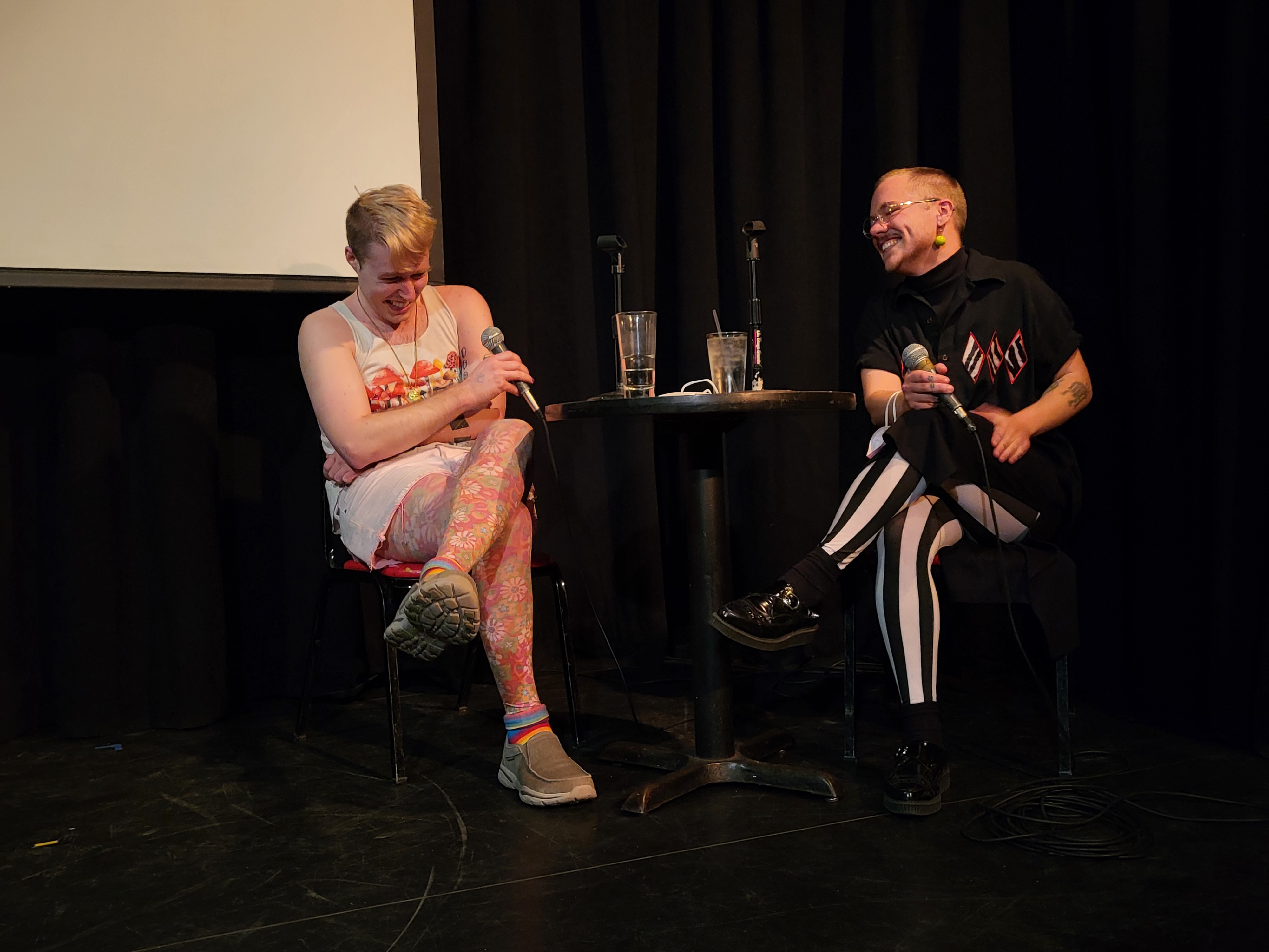 MFA Mentor Image Merit Thursday, two people laughing with microphones on a dark stage