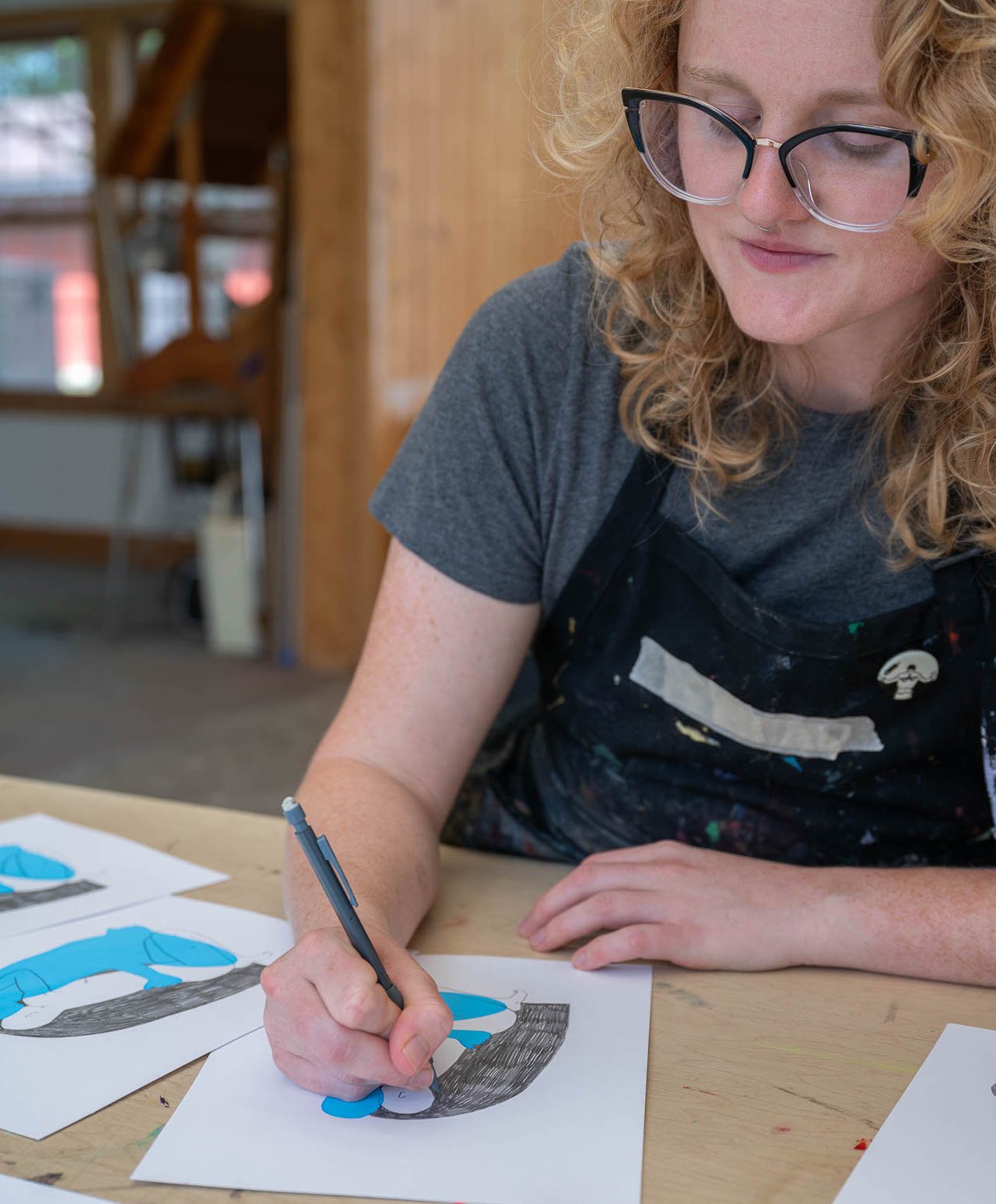 MFA Mentor Image Sarah Everson, person with glasses, wearing an art smock, drawing in an art studio