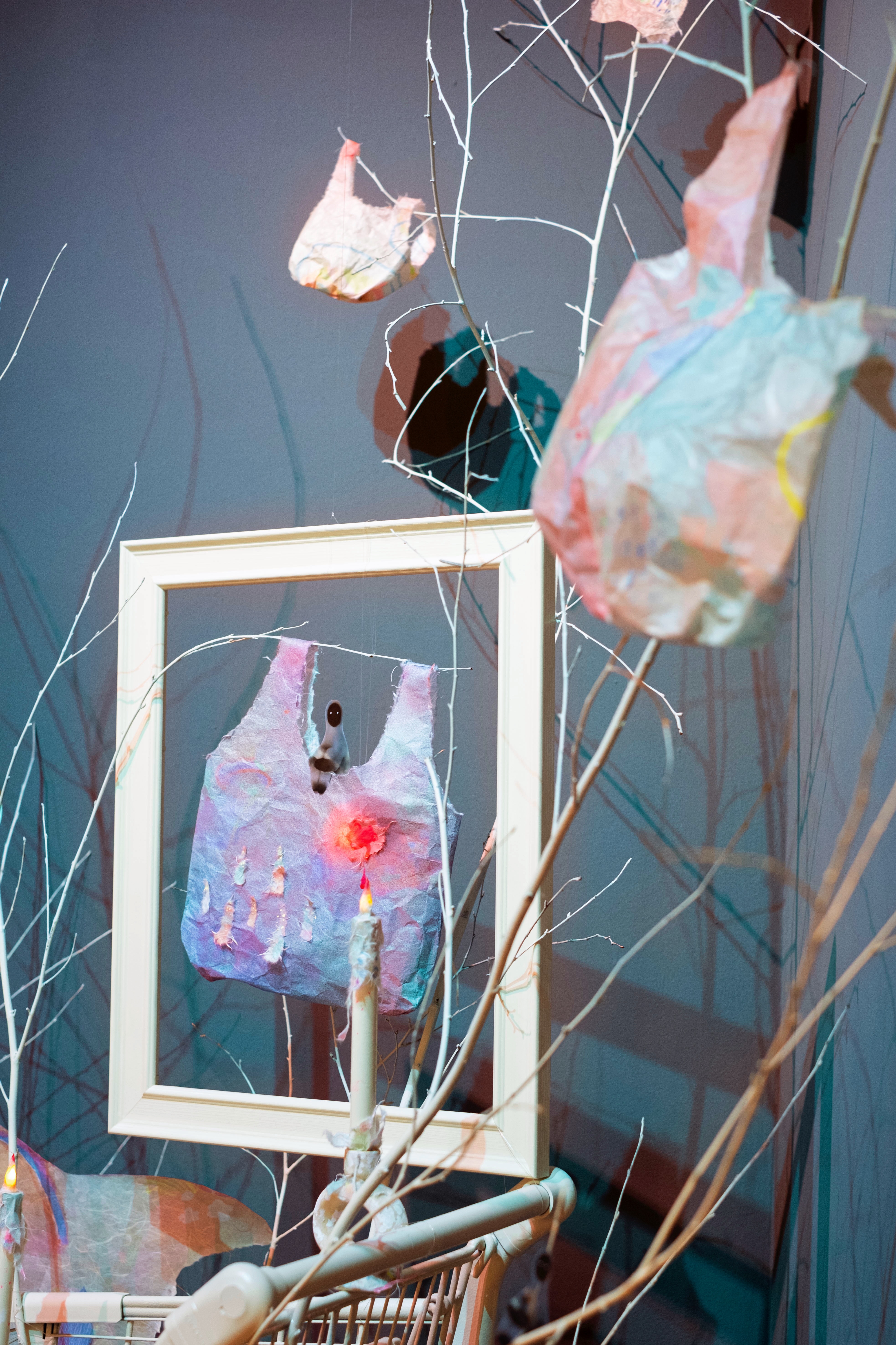 Glimpse of Yi's installation featuring plastic bag form in a frame, thin tree branches, candle and shopping cart