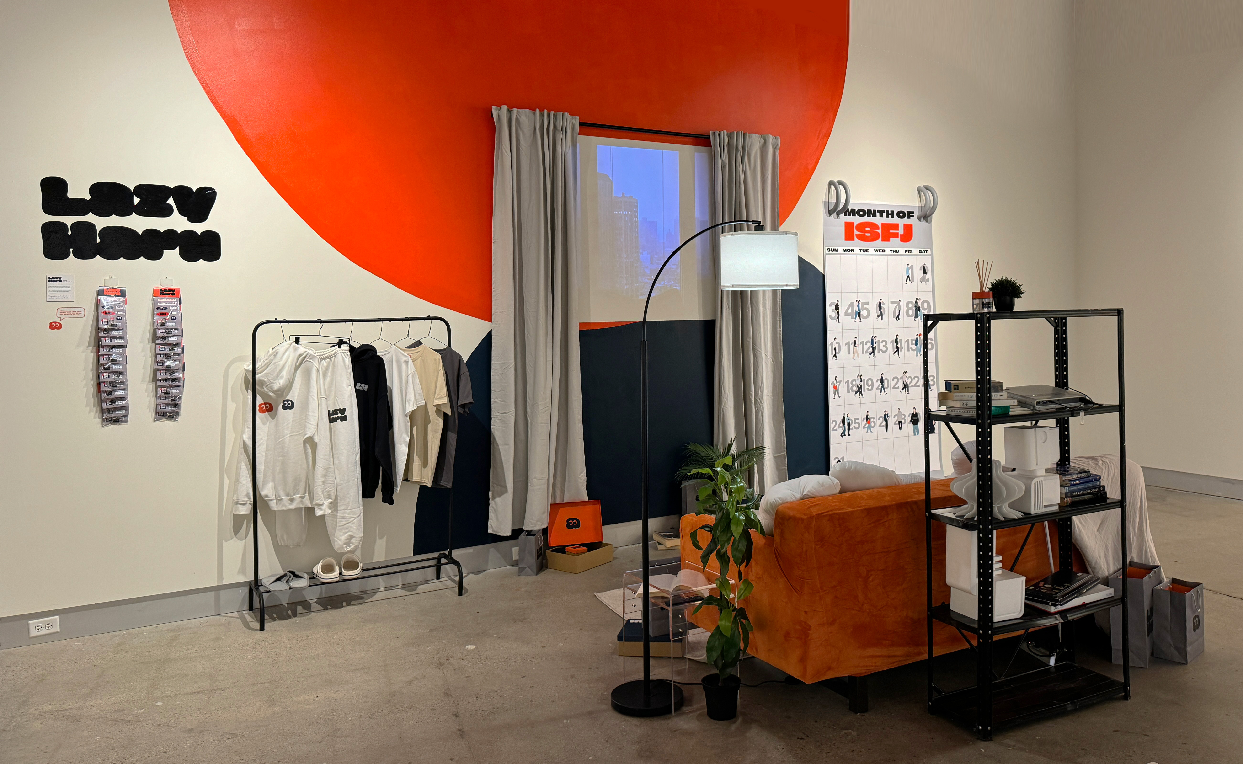 Image of Ria's thesis work. A modern room setup with a bold red accent wall. Clothes hang on a rack, a large floor lamp arcs over an orange couch, and a shelving unit holds various items, including books and electronics. A large window shows a cityscape, and a calendar titled 'MONTH OF ISFJ' is on the right.