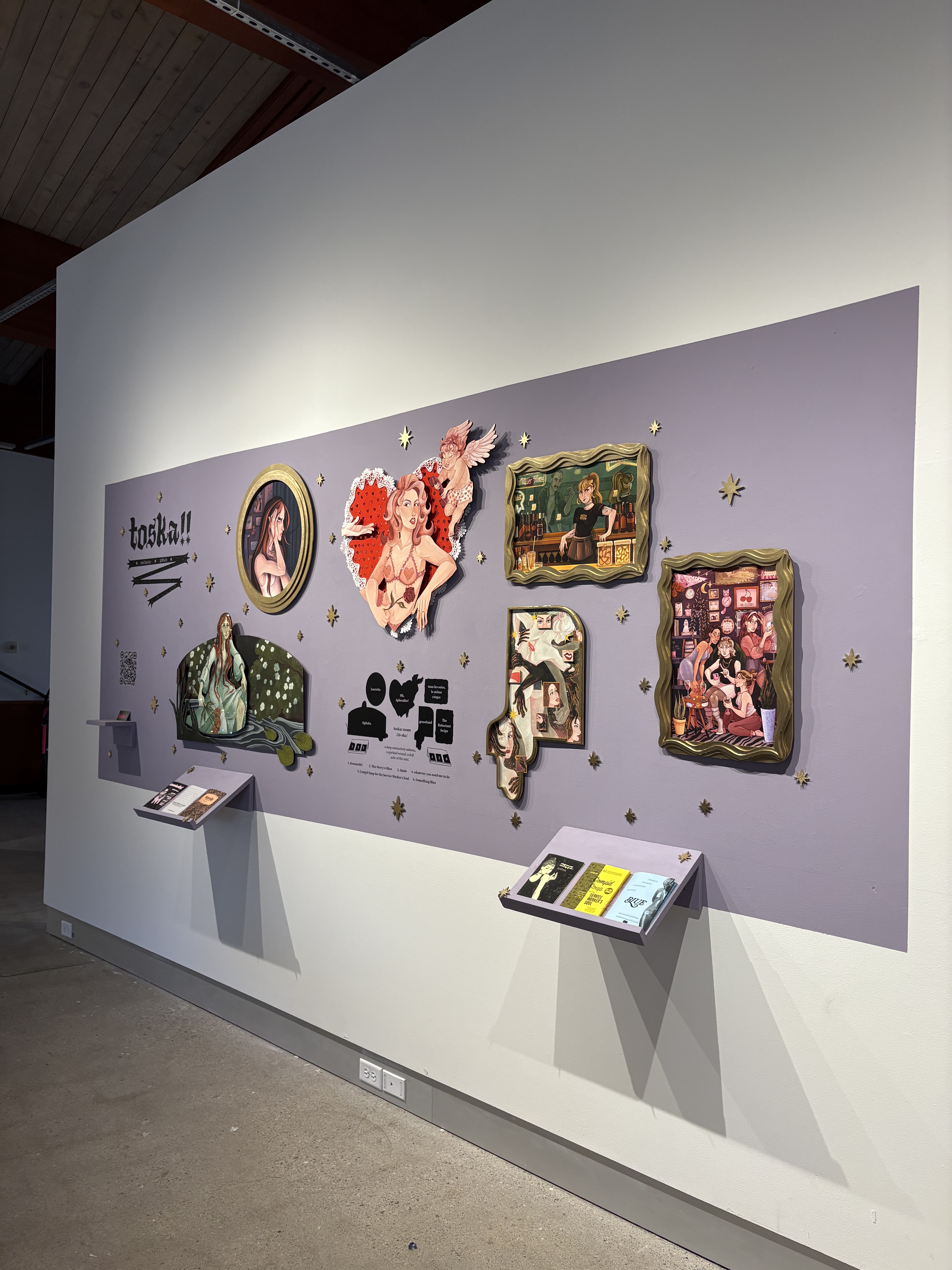Image of Michayla‘s work. An art exhibition wall with colorful framed illustrations, a lavender backdrop, and two display shelves holding booklets