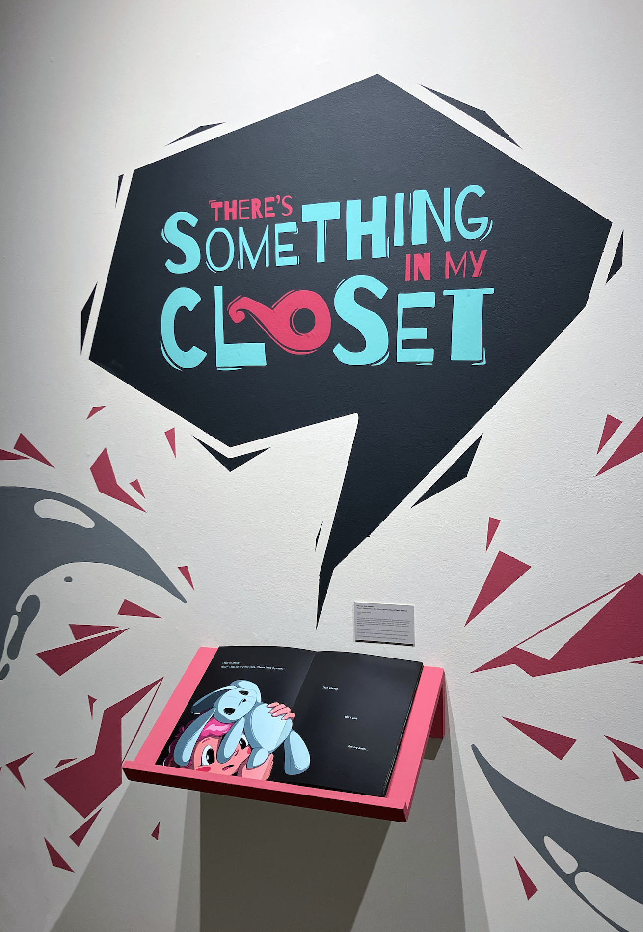 Image of Miranda Hoover's work. Wall text reads 'There's Something in My Closet' above an open illustrated book on a pink shelf."