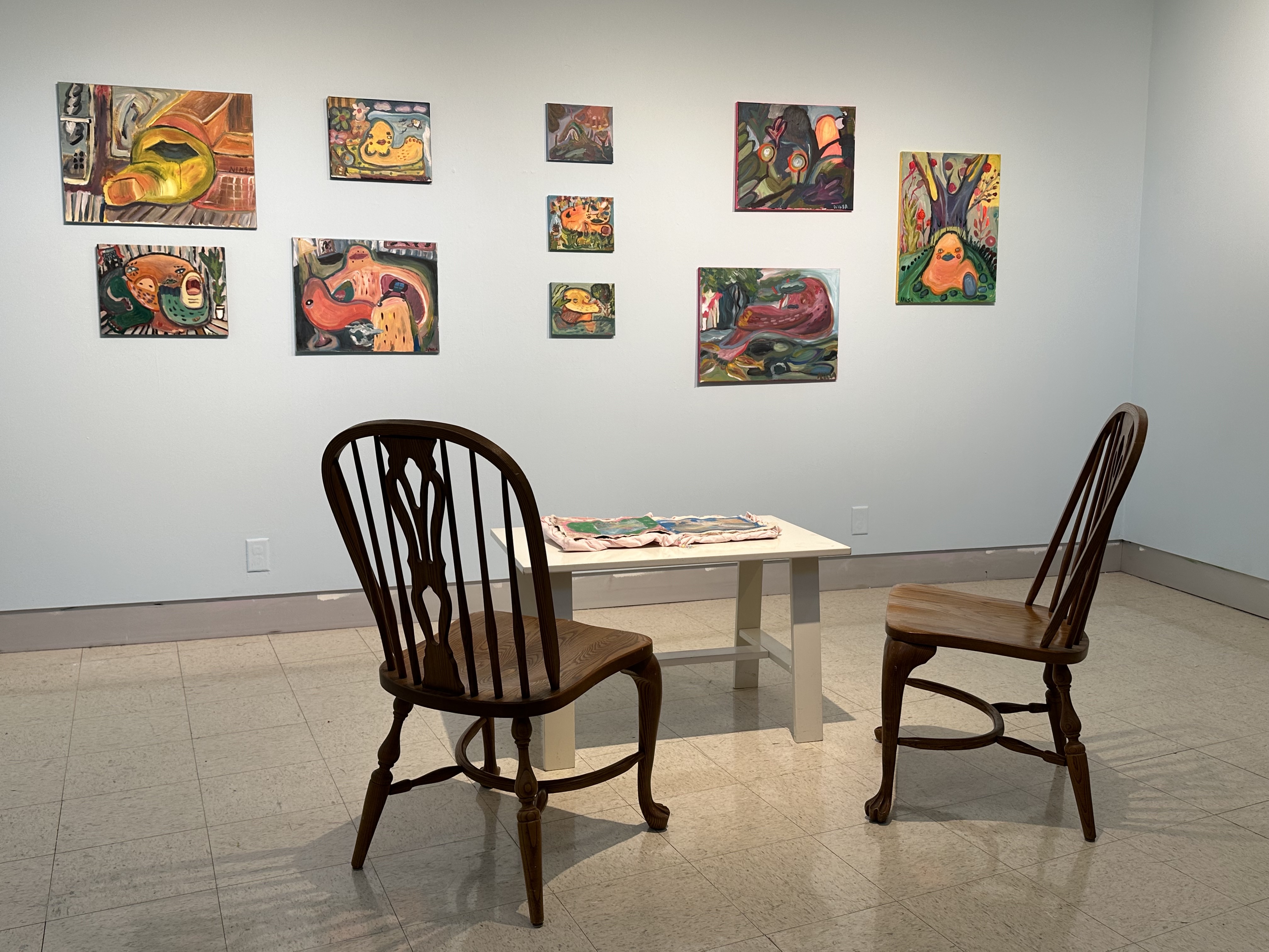 Installation of paintings on a wall. And a table with a handmade book, chairs to sit in.