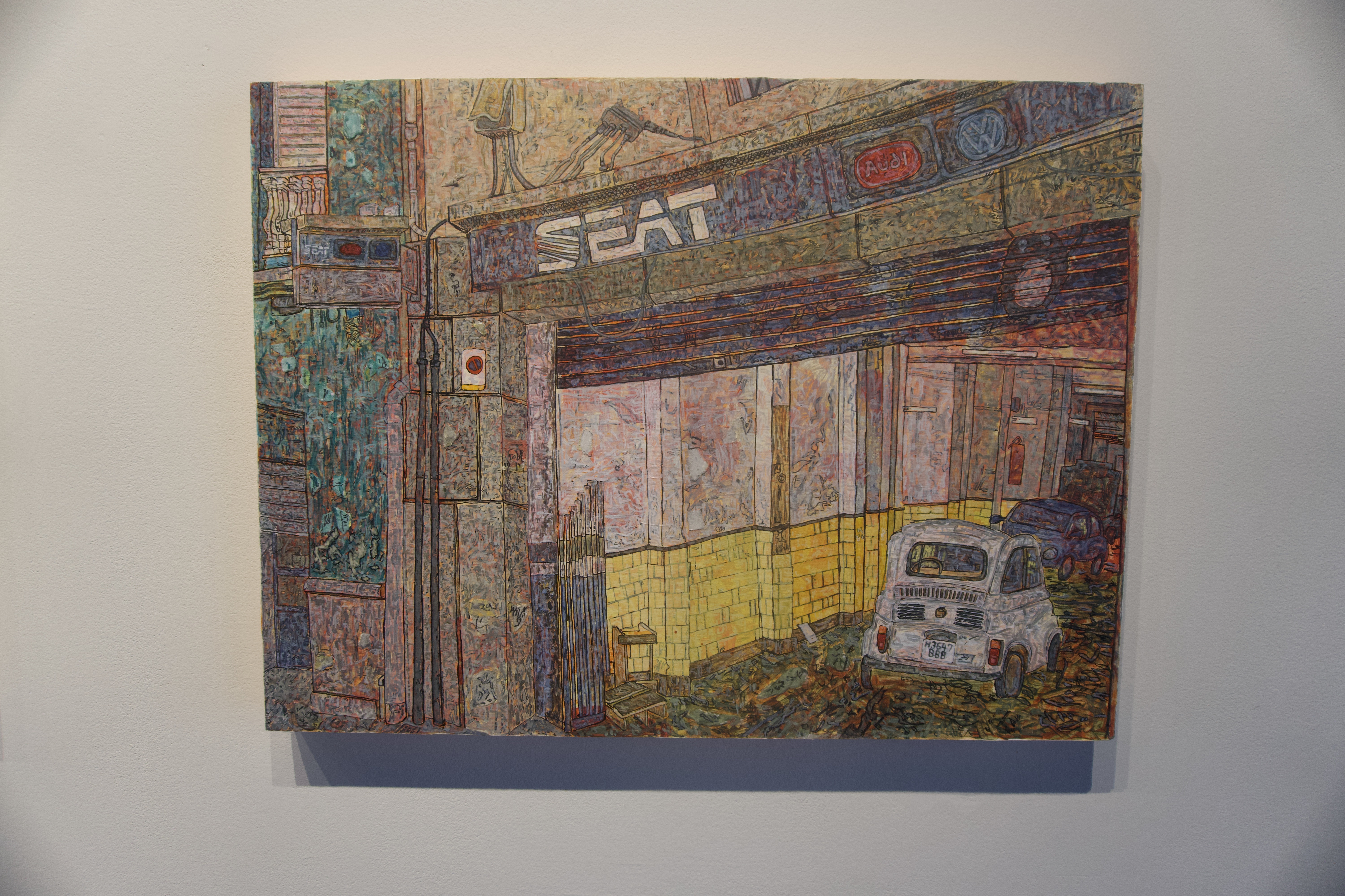 Image of Matthew Clara's work. A textured painting of an old car repair shop with SEAT, Audi, and Volkswagen logos above the entrance. The scene shows a small white vintage car parked inside, surrounded by worn yellow tiles and weathered walls. The artwork is rich with intricate, colorful brushstrokes, creating a layered, almost mosaic-like effect."