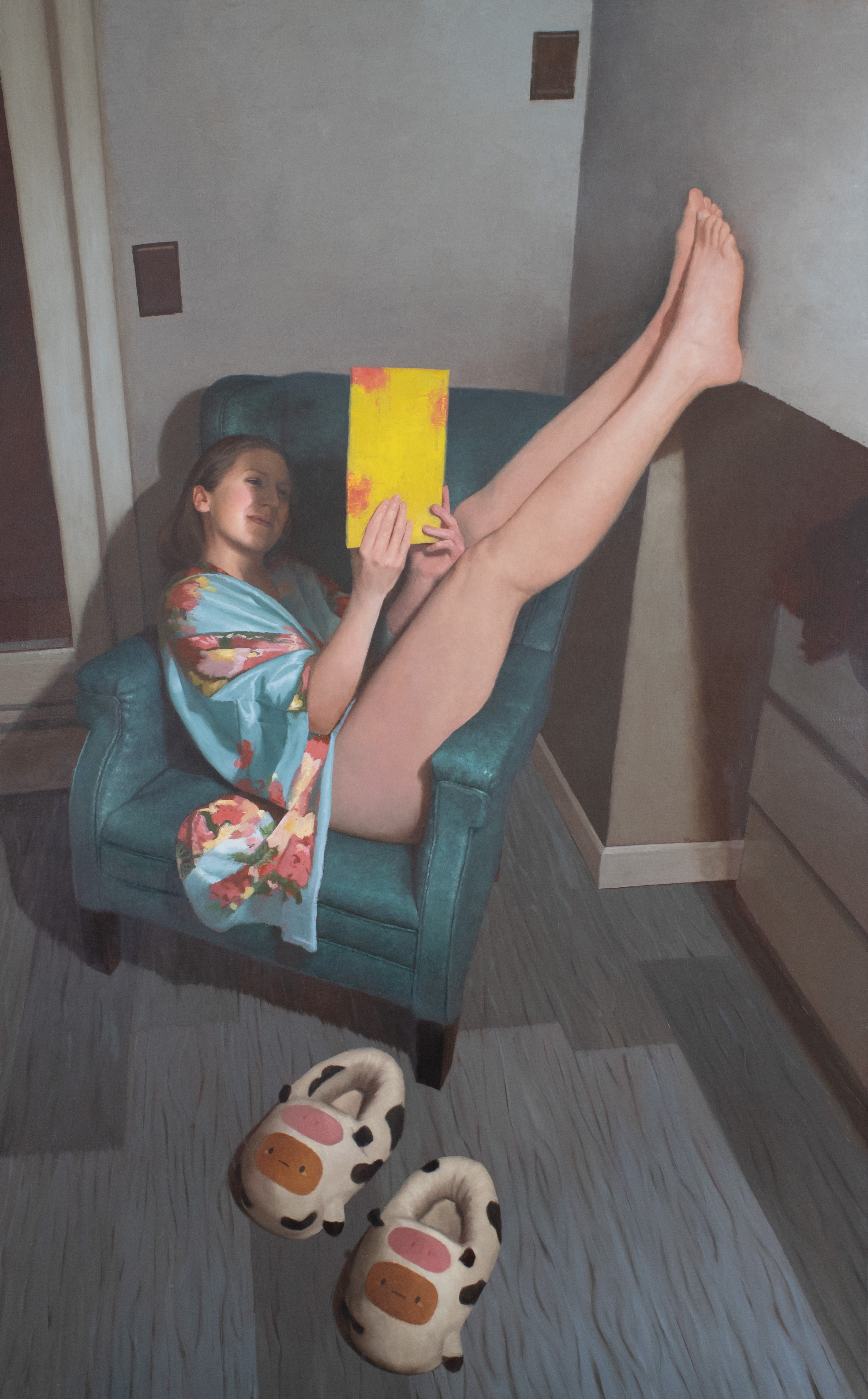 Person in robe reclines in chair reading a yellow book, smiling, with plush cow slippers in the foreground