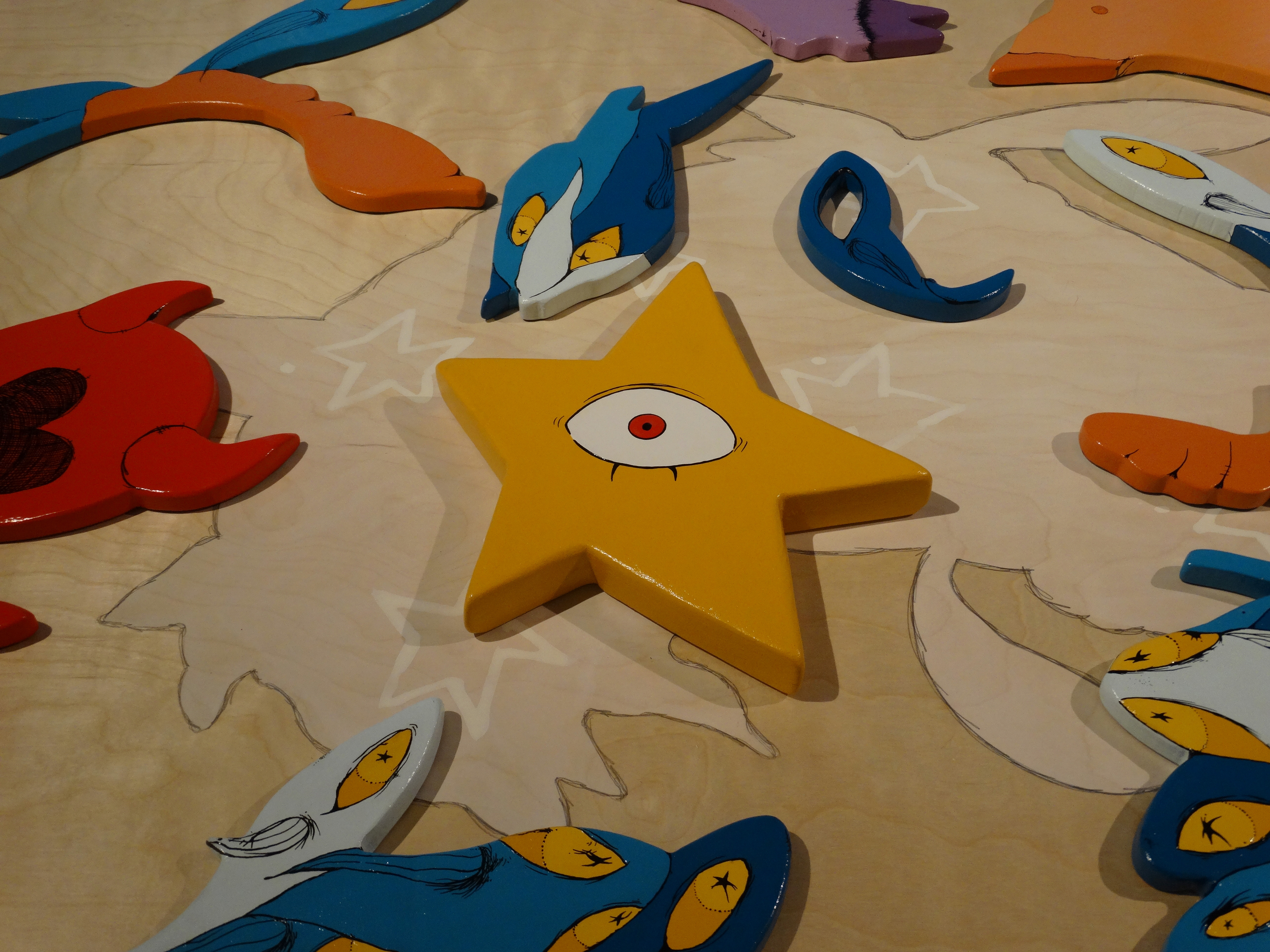 Image of Elizabeth Blackwell's work. Wooden painted pieces, including a yellow star with an eye, are arranged on a light wood surface with animal-shaped outlines and colorful abstract forms.