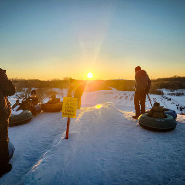 People are sledding down a snowy hill, there is a yellow sign that reads "WAIT TO GO UNTIL STAFF SIGNALS"