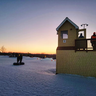 A small yellow building is on a snowy landscape, and on it there are two people. People are sledding in the background