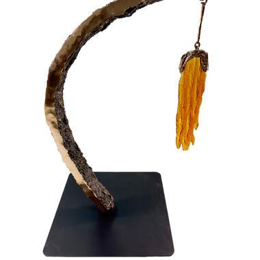 Artwork by John Hallett: Abstract sculpture with a square, metal base, bronze vertical projection arcing over the base, and orange glass hanging from the end of the bronze