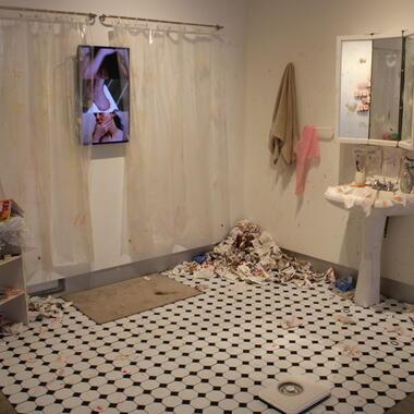 Artwork by Elsie Gray: an installation of a tiled bathroom floor, pedestal sink and mirror, overflowing wastebasket. A small television on the wall plays a video of the artist.