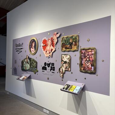 Image of Michayla‘s work. An art exhibition wall with colorful framed illustrations, a lavender backdrop, and two display shelves holding booklets