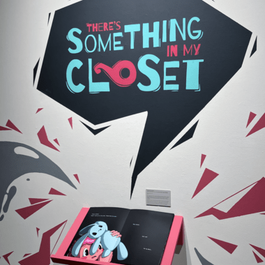 Image of Miranda Hoover's work. Wall text reads 'There's Something in My Closet' above an open illustrated book on a pink shelf."