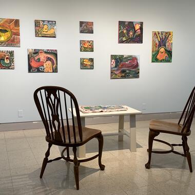Installation of paintings on a wall. And a table with a handmade book, chairs to sit in.