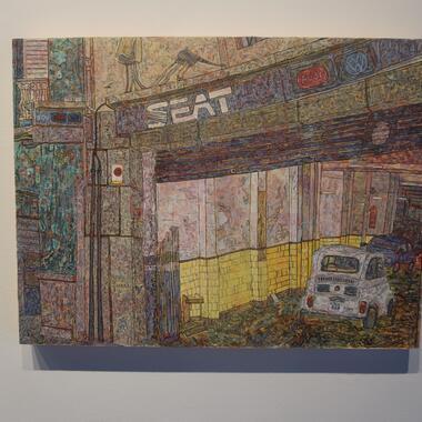 Image of Matthew Clara's work. A textured painting of an old car repair shop with SEAT, Audi, and Volkswagen logos above the entrance. The scene shows a small white vintage car parked inside, surrounded by worn yellow tiles and weathered walls. The artwork is rich with intricate, colorful brushstrokes, creating a layered, almost mosaic-like effect."