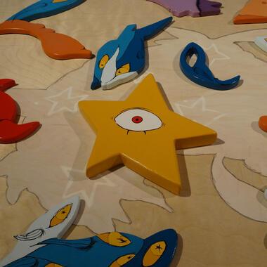 Image of Elizabeth Blackwell's work. Wooden painted pieces, including a yellow star with an eye, are arranged on a light wood surface with animal-shaped outlines and colorful abstract forms.