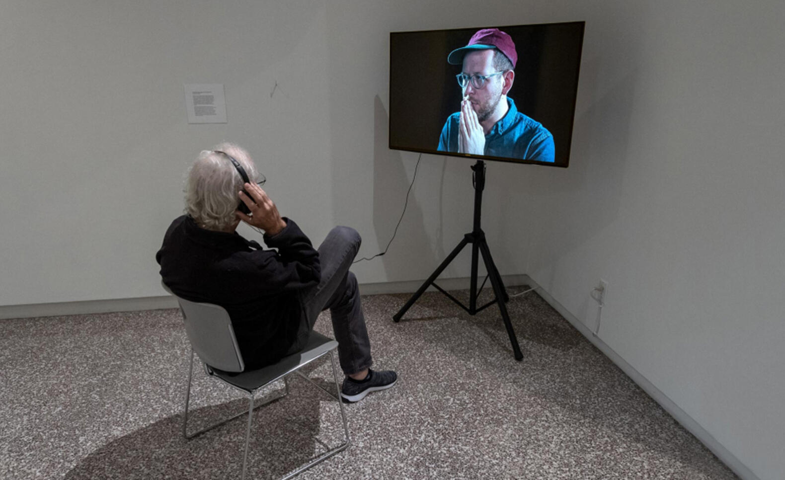Man sitting in a chair looking at a screen which displays another man holding hands over his mouth ; Gonzalo Reyes Rodriguez