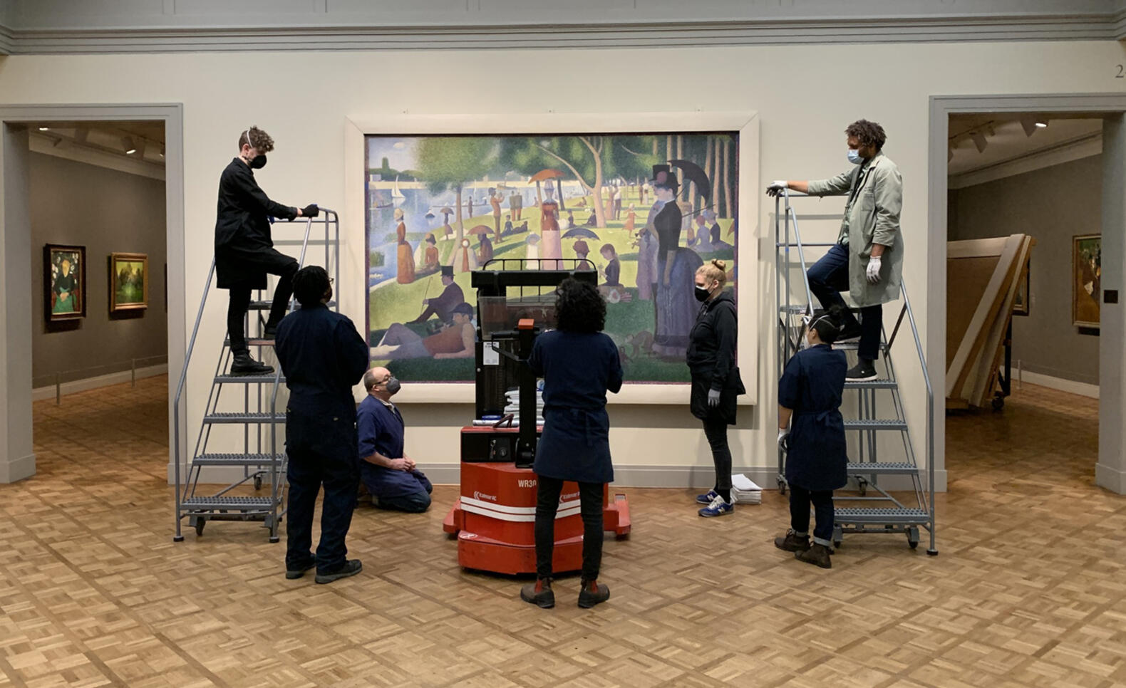Several people gathered around the painting "A Sunday Afternoon on the Island of La Grande Jatte" all putting a frame onto it