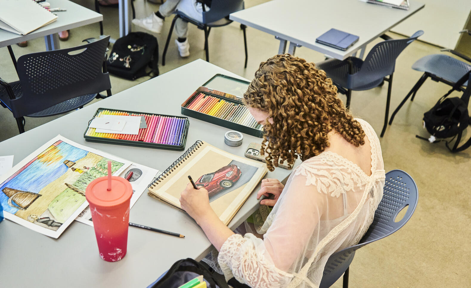 Student works at a table on colorful drawings in colored pencil with two colored pencil sets on the table.