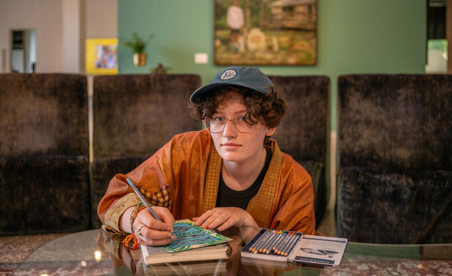 Student sits at a shiny table, looking directly at camera, as they work on a sketchbook. The student is wearing an orange shirt and blue baseball cap. The background shows a green wall and abstract image.