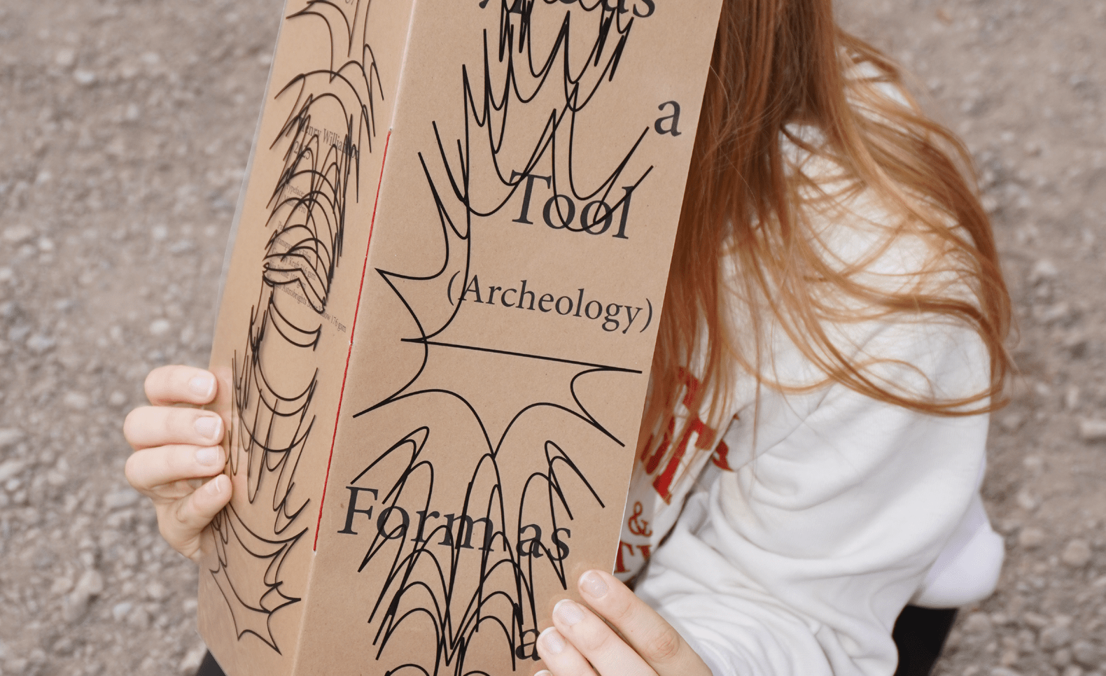 A blonde person kneel with their face covered by a pamphlet titled Text as a Tool, Form as Accident. ; Henry Williamson