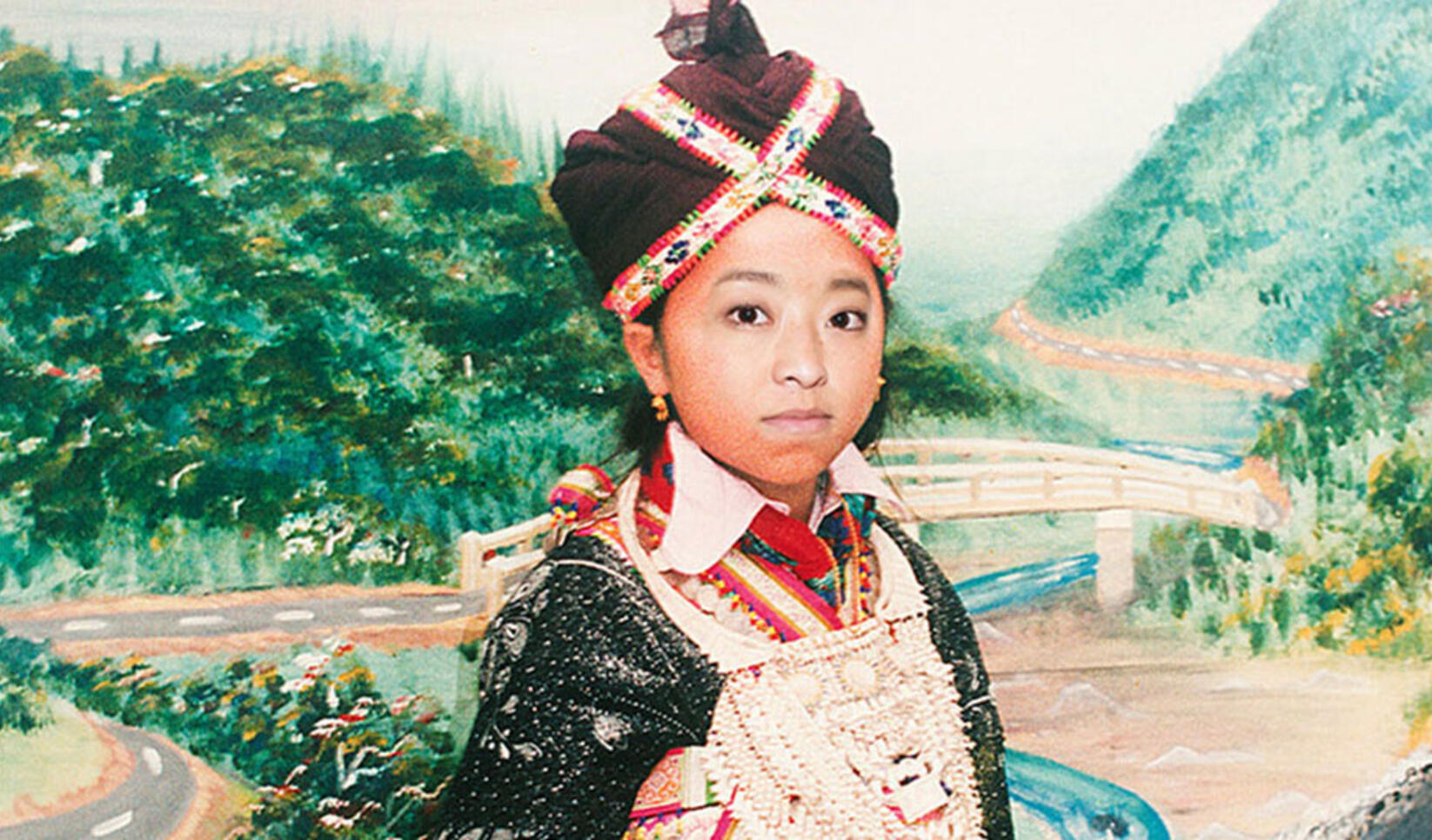 Young Girl stands infront of a large painting background dressed in traditional Hmong attire. ; Pao Her