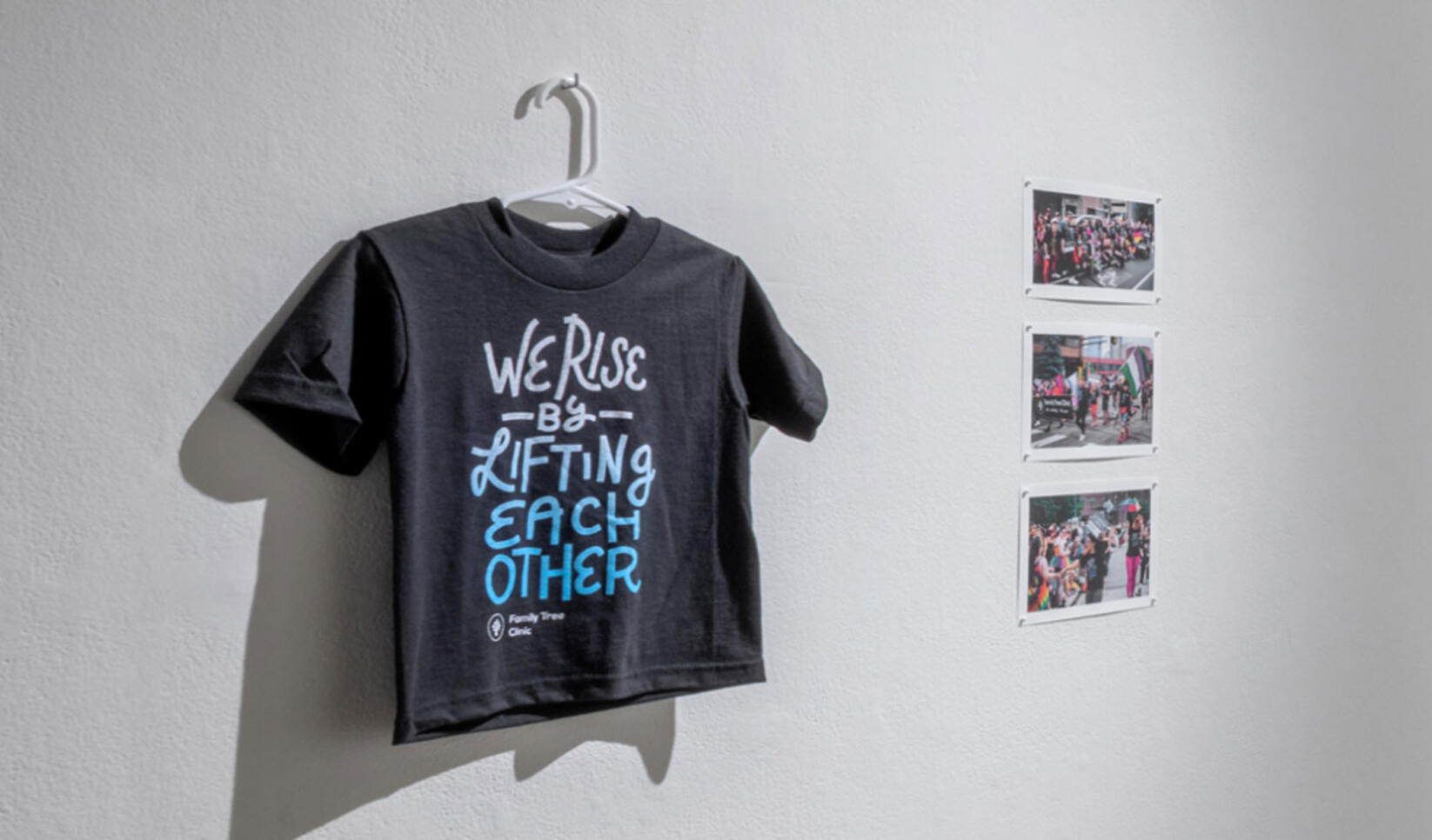 Shirt with multicolored text on it reading "We Rise by Lifting Eachother" ; Alison Nowak