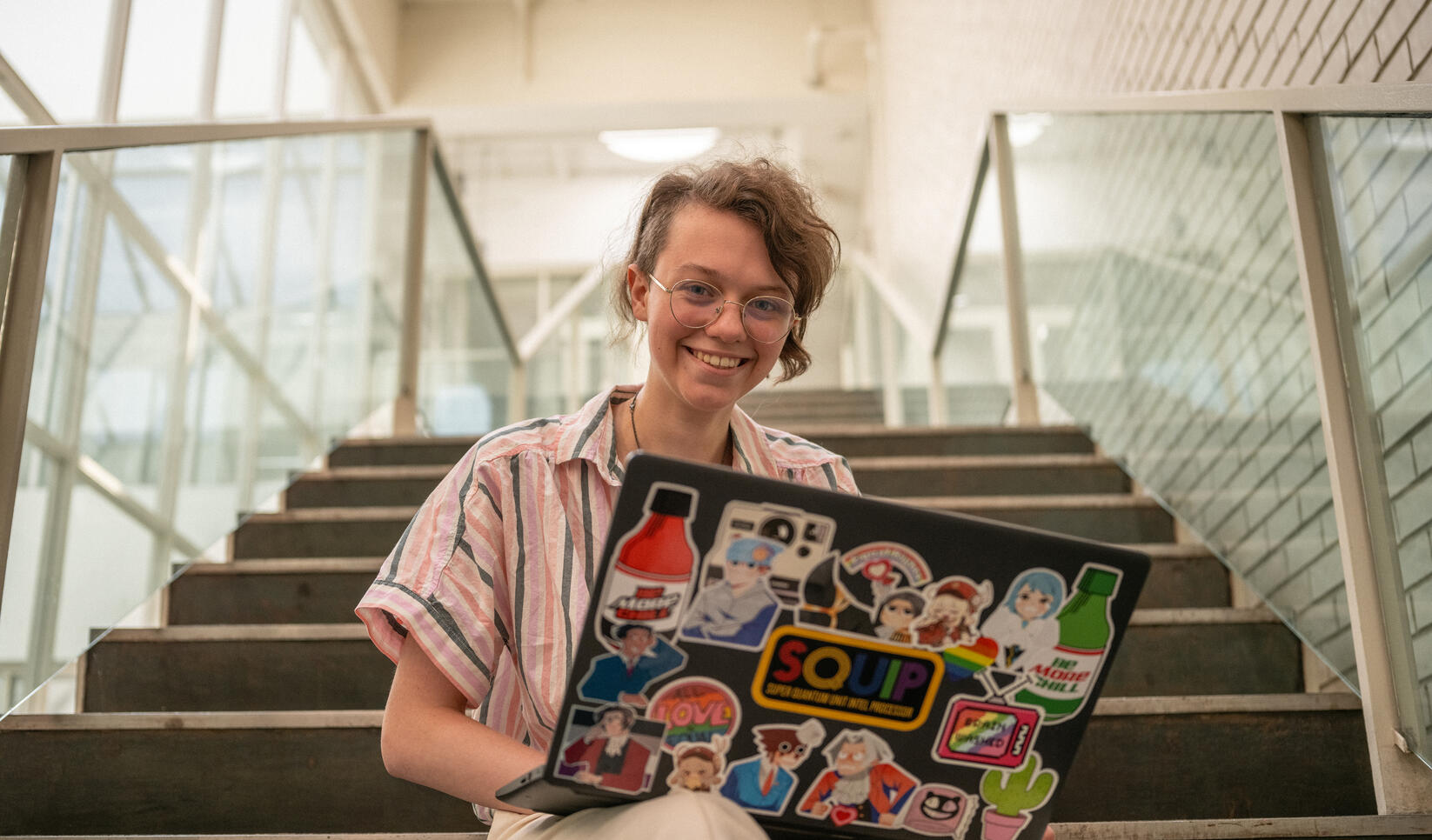Student sits on stairs looking into camera and smiling with laptop in lap. The camera looks up on the stairs and several colorful stickers can be seen on the laptop.