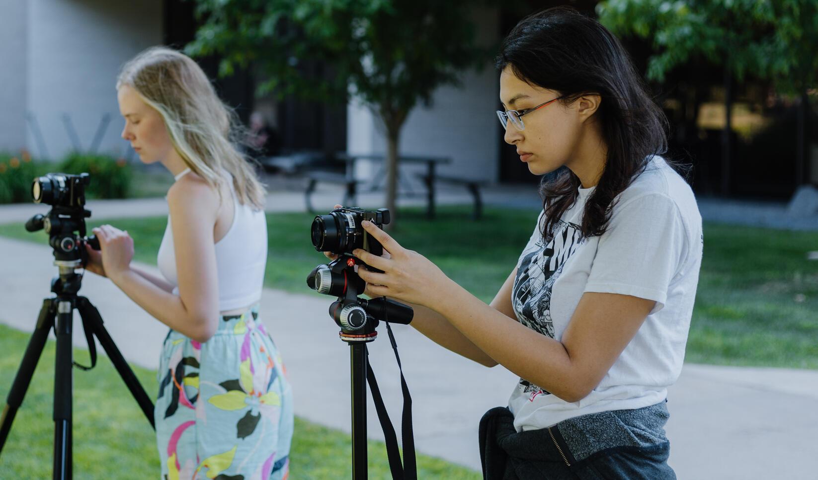 Two students stand outside with their cameras on tripods. 