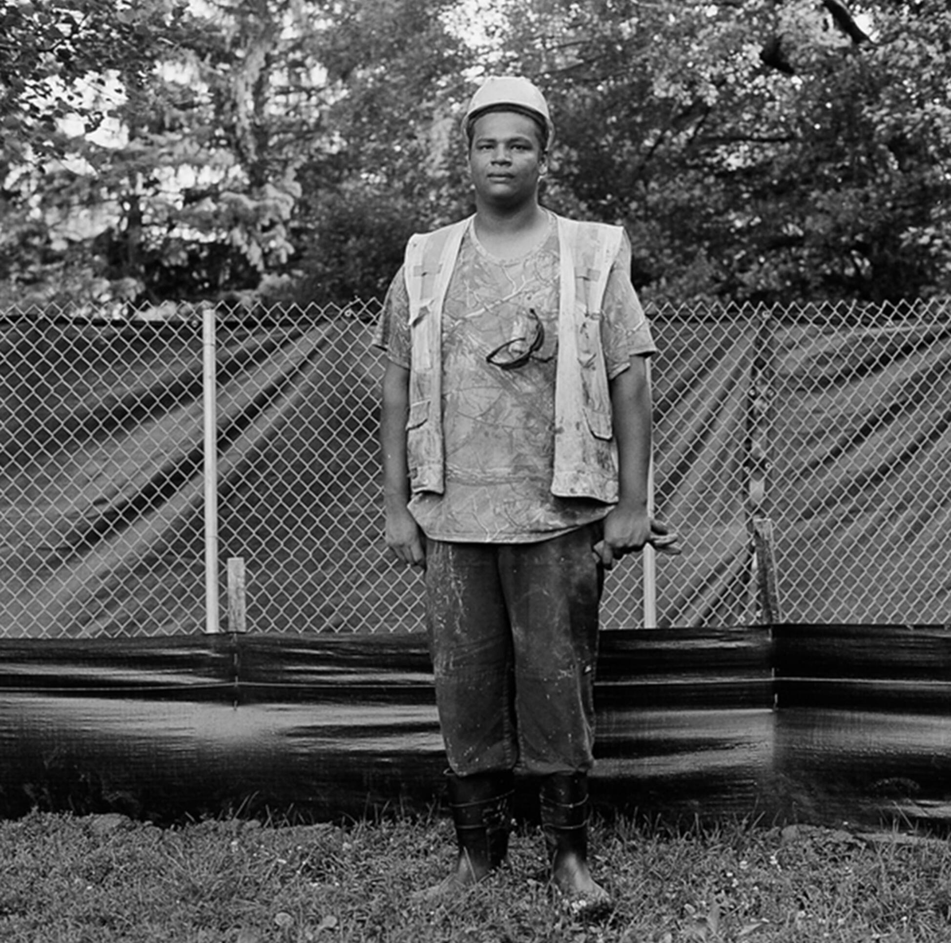 Black and white photograph of a young man in a hard hat in front of a chained fence ; Leslie Grant