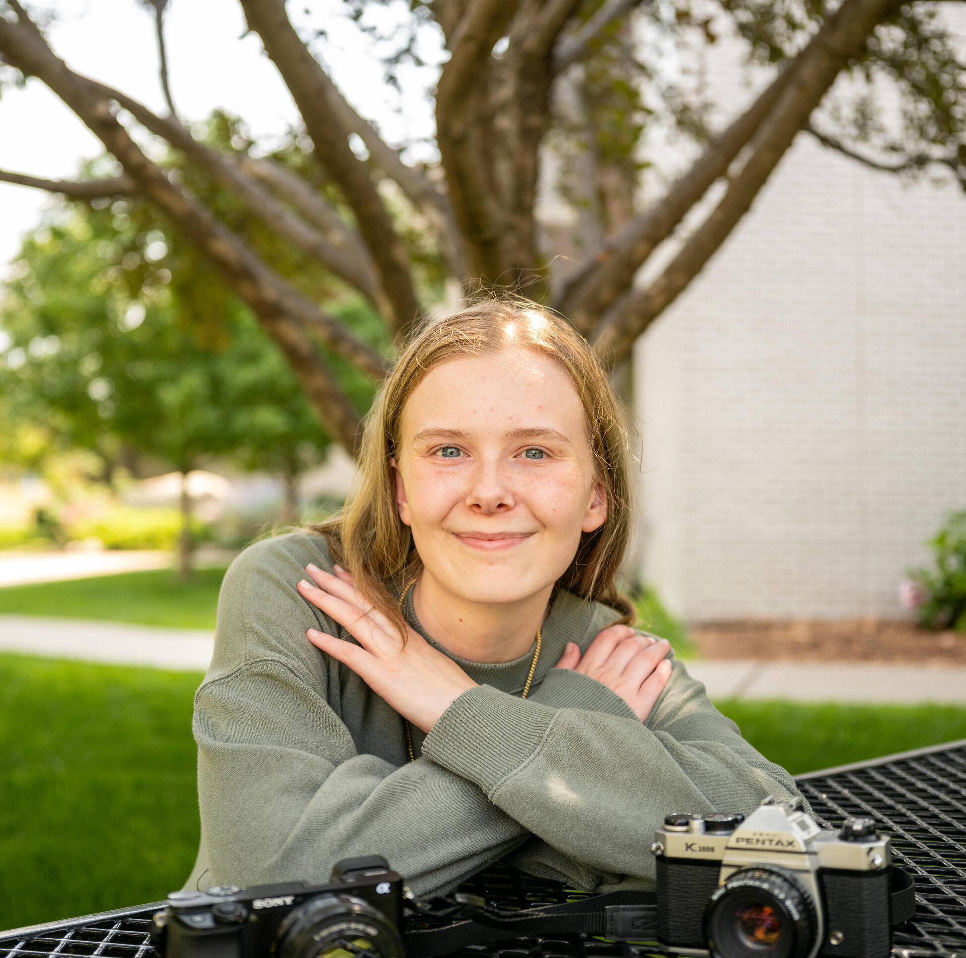 Student sits at picnic table outside of MCAD looking directly into the camera with two cameras on the table in front of her.