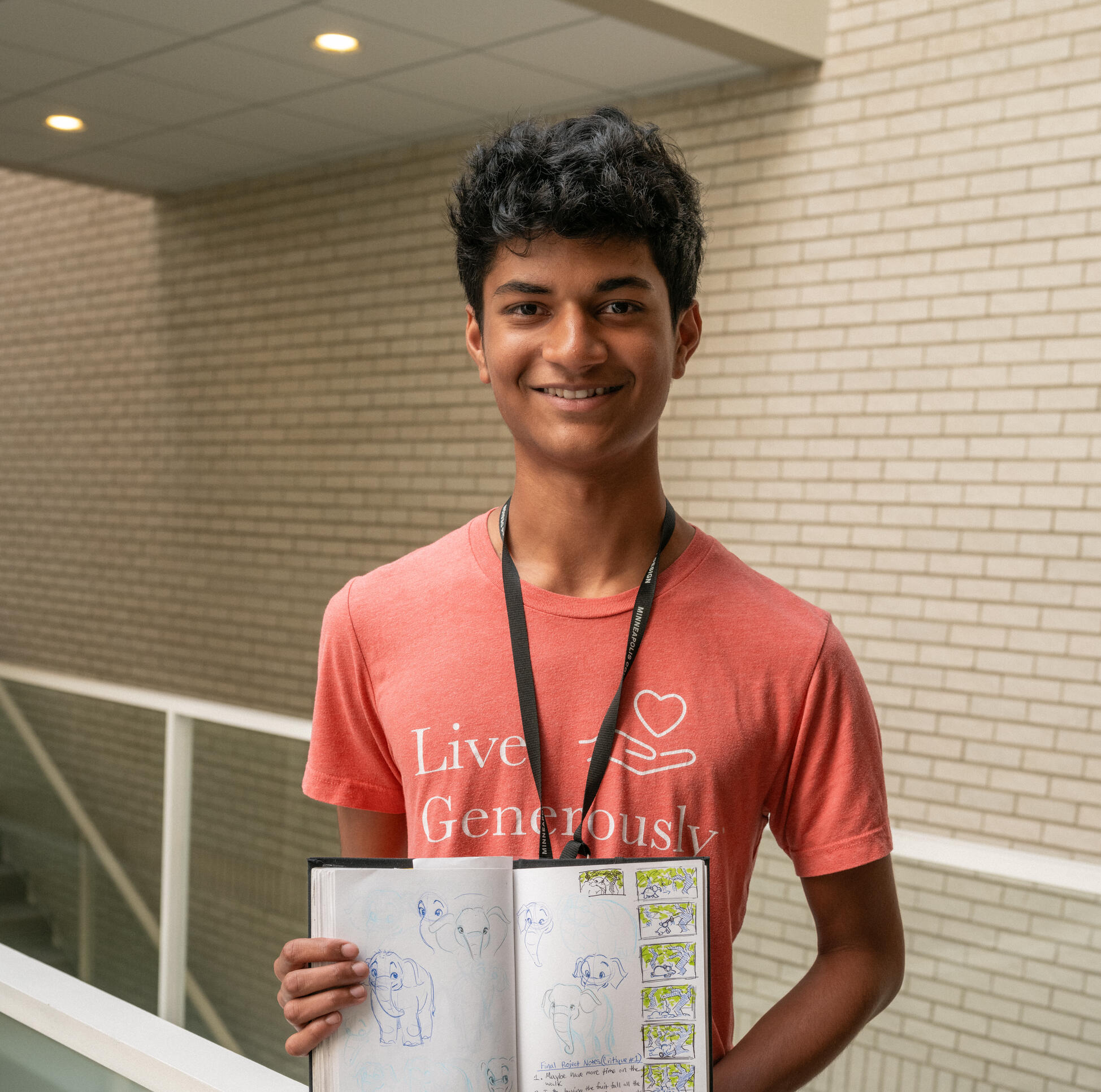 Student stands on the MCAD bridge (inside of MCAD), holding their sketchbook open so the audience can view. Student wears a red t-shirt and looks directly at the camera.