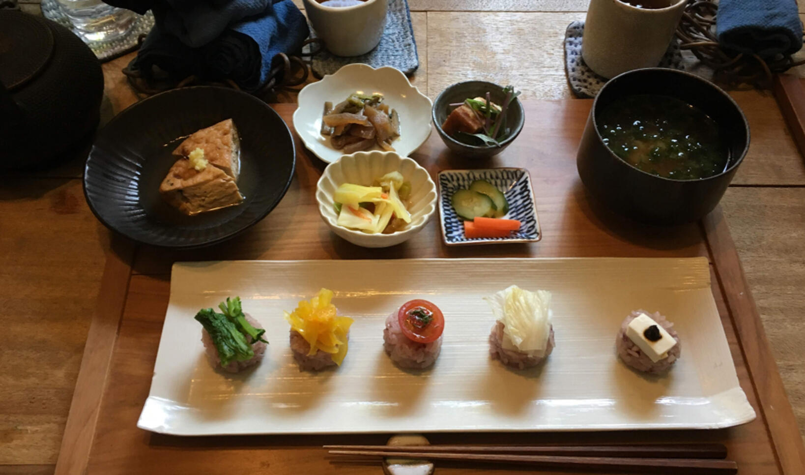 Several colorful dishes laid out on individual plates and all laying on a wooden board ; Aki Shibata
