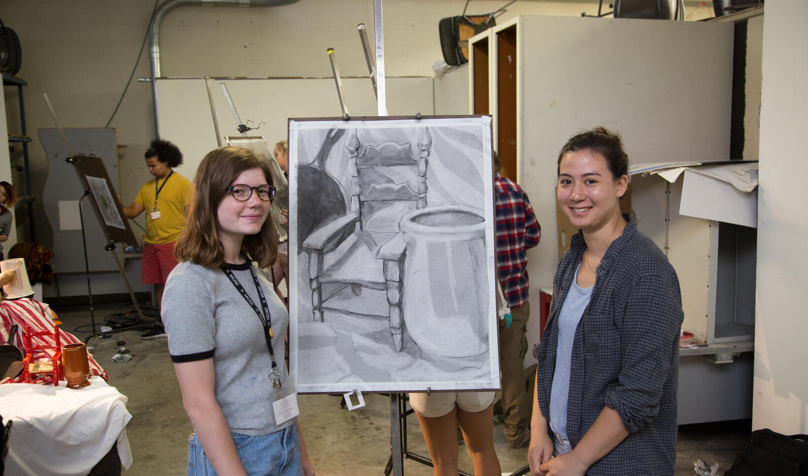 Two young artists stand on either side of a black and white drawing of a still life in the drawing studio.