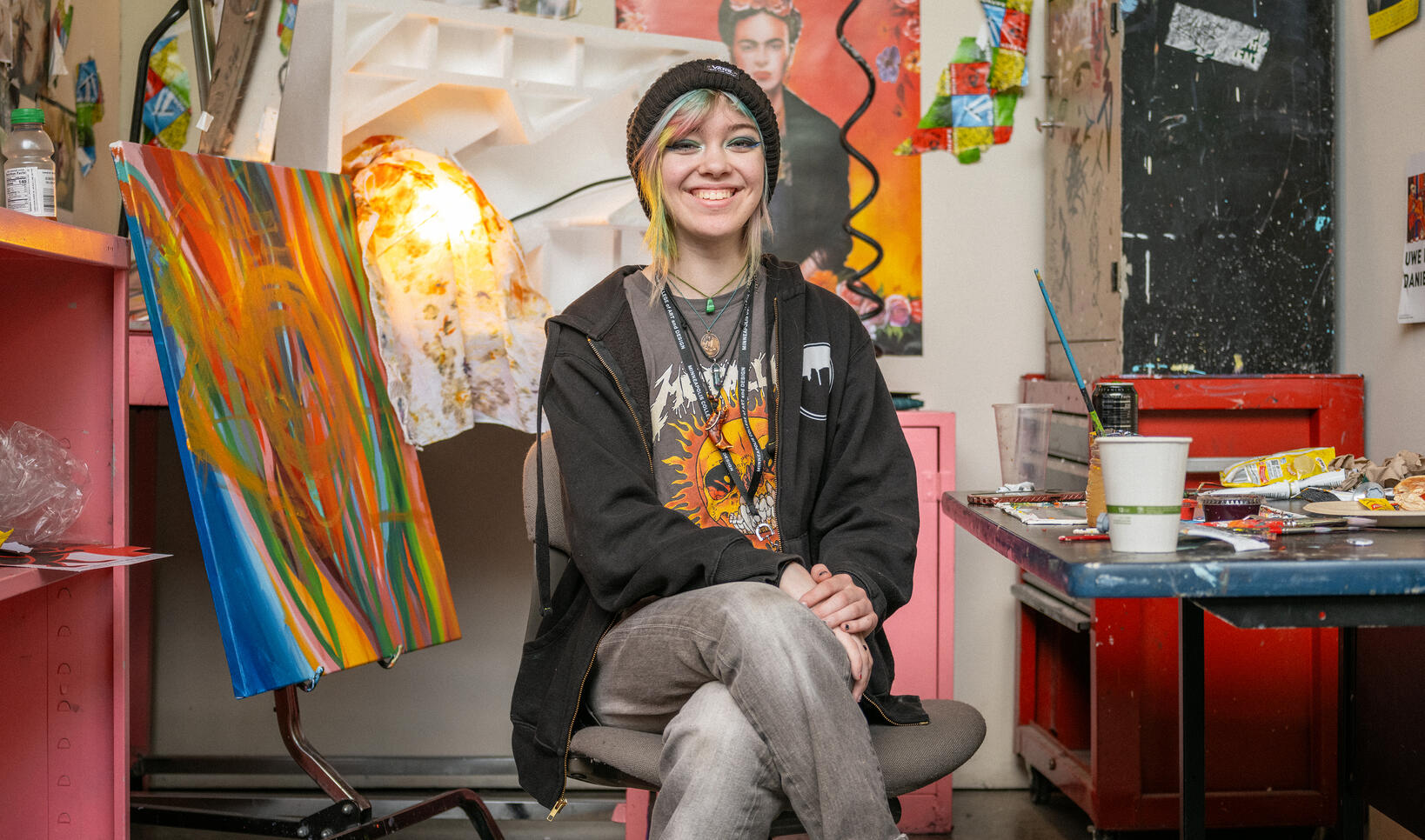 Student sits on a chair facing the camera in a painting studio with several brightly colored paintings and posters on walls.