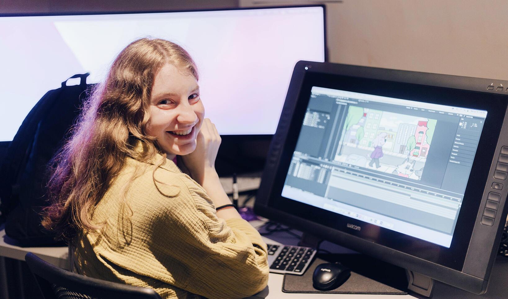 Student sits at an animation computer with an animation still showing on her computer. Student looks over shoulder at camera and smiles. Her backpack can be seen in the background.