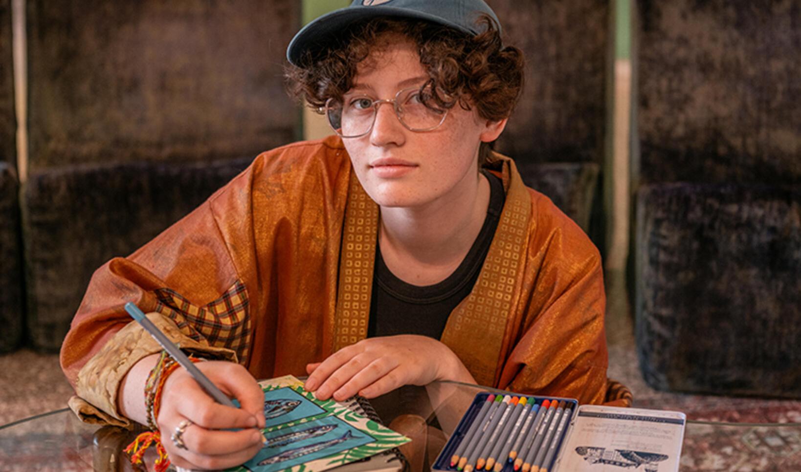 Student sits at a shiny table, looking directly at camera, as they work on a sketchbook. The student is wearing an orange shirt and blue baseball cap. 