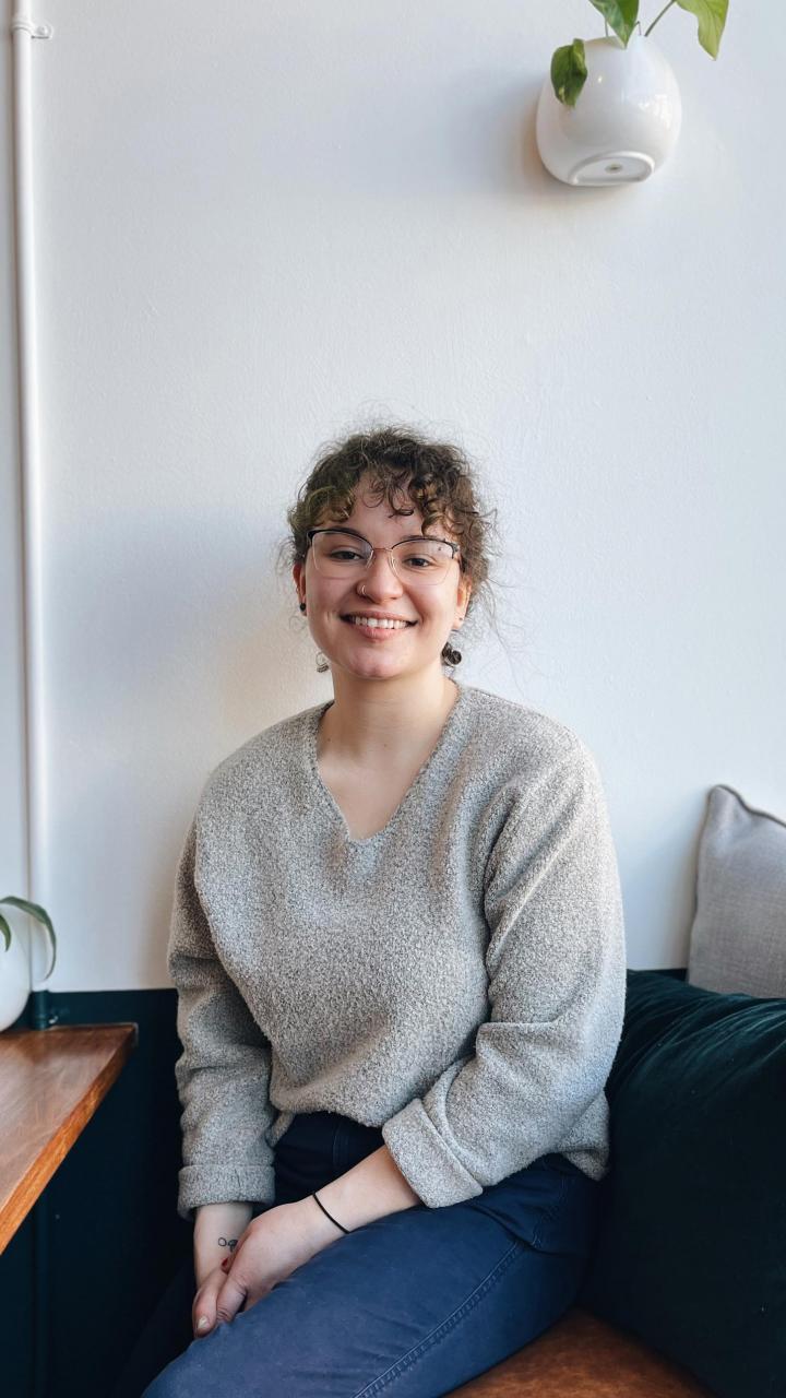 Portrait of Sarah Hormanski. She is sitting at a desk smiling with a gray shirt and jeans.