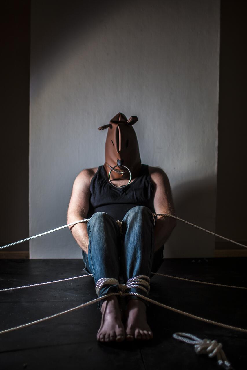 Photograph of a figure roped down in to sitting position while wearing a sculptural mask.