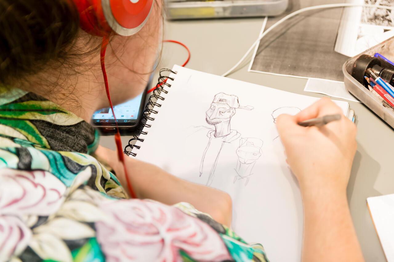 Student seen from behind drawing a character on a notebook with pencil.