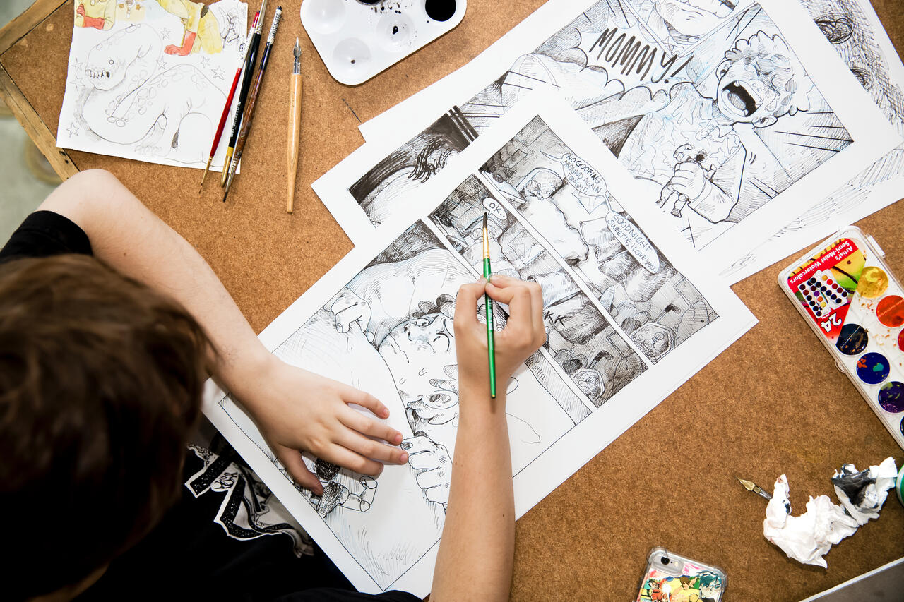 Looking overhead at a student drawing comics on a wood table with supplies on either side of the paper.