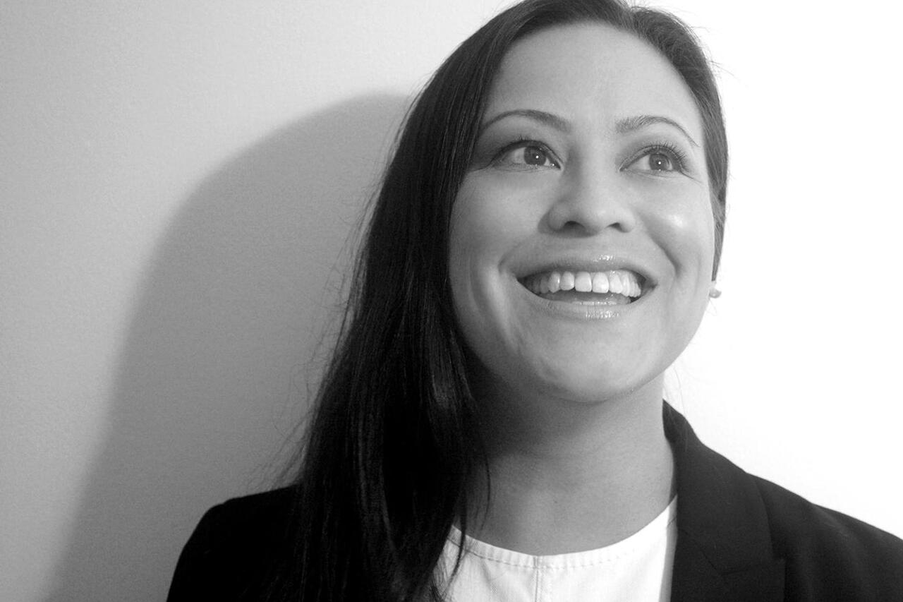 A black and white photograph of a woman smiling looking upwards towards the right. The woman is wearing a blazer and white shirt and she has long dark hair.