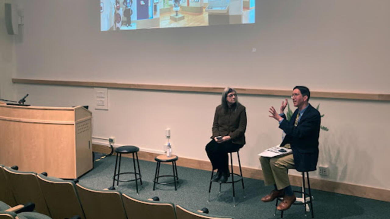 Two people sit in front of a projector and they are both in high stools