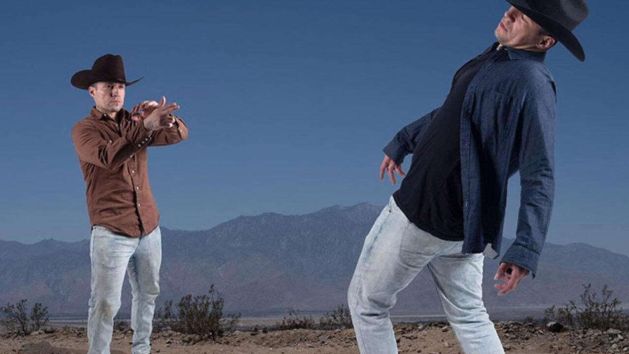 Two men are standing in cowboy hats, the man on the left is pointing his fingers in the shape of a gun at the man on the right who is leaning back as if he was shot