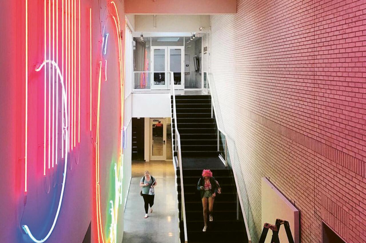 Two people walking in MCAD's Main Building with a neon installation on the wall