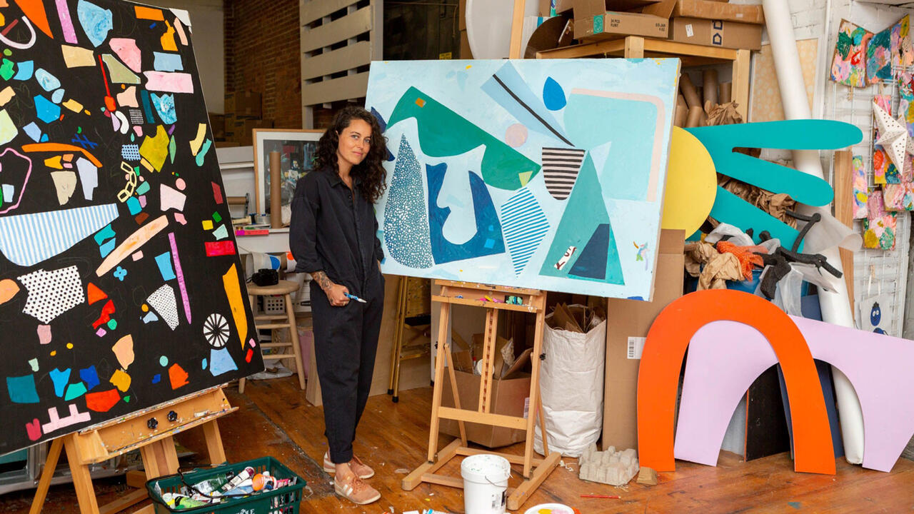 A woman dressed in all black stands with many colorful paintings on easels