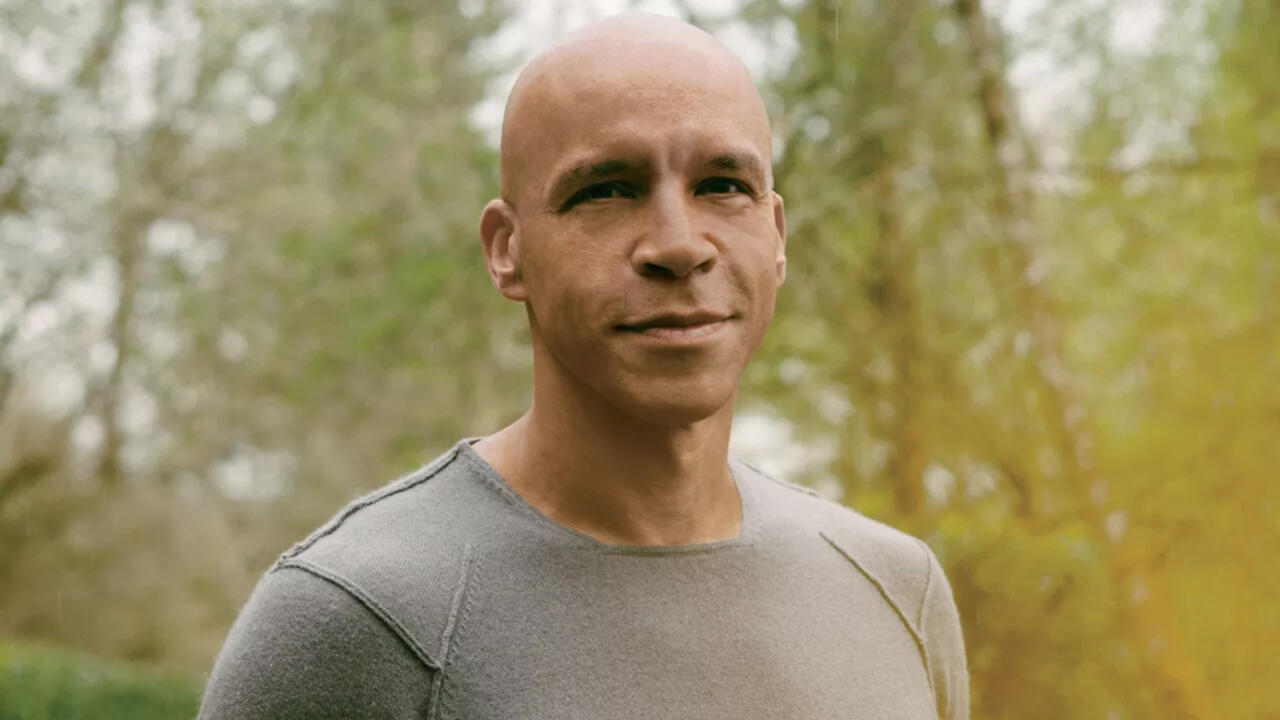A man wearing a tight gray shirt stands in-front of green trees and forest background smiling at the camera