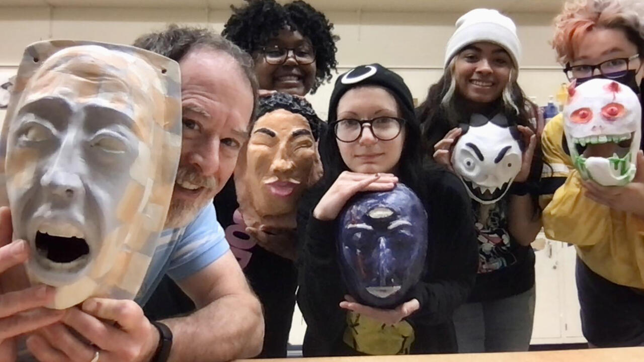 a middle aged man sits in-front of a group of young adults all holding different hand painted masks