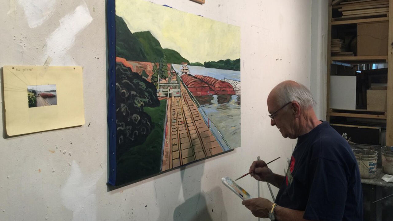 An older man is sitting in front of a painting of a bridge and body of water, he is in the middle of painting and is looking down at his palette